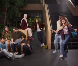 Cinematisch geënsceneerde groepsfoto in een universiteitsgebouw met trap en zitplekken, waarin studenten elkaar ontmoeten, samenwerken en zich door de ruimte bewegen. Groep studenten in een modern atrium: vier zitten met laptop op houten bank, twee staan bij trap en één stel omhelst elkaar.