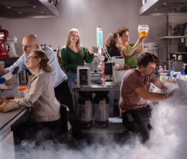 Cinematisch geënsceneerde foto van toegepast onderzoek in een labkeuken, waar experimenten met voedsel en meetapparatuur samenkomen in een toekomstgerichte werkomgeving. Vijf mensen werken in een labkeuken met microscopen en glazen potten; op de voorgrond ligt brood en hangt er witte rook.