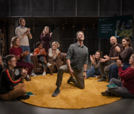 Geënsceneerde groepsfoto in een onderwijs- of trainingsomgeving waarin een groep volwassenen een centraal moment van erkenning en verbondenheid markeert. Groep mensen zit en staat in een cirkel op een gele vloerkleed; in het midden knielt één persoon terwijl de anderen applaudisseren.